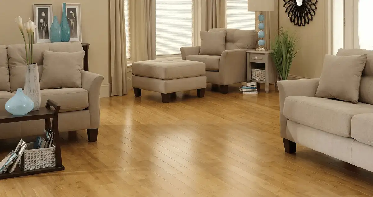 A living room with professionally installed bamboo flooring, beige sofas, a matching ottoman, a side table with a blue lamp, vases, framed pictures, and large windows with blinds and curtains in Boise, ID